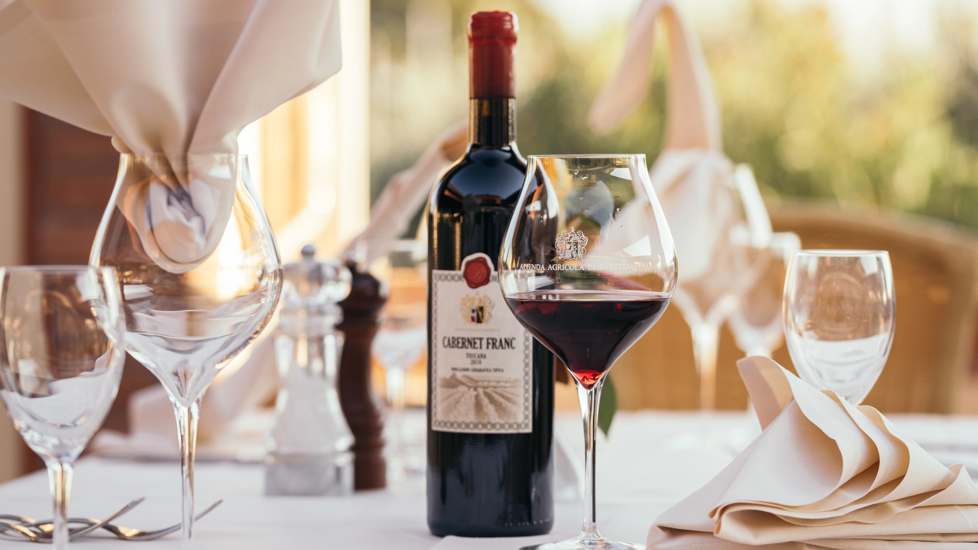 Un calice di vino rosso e una bottiglia di Cabernet Franc prodotto dalla Tenuta Elisabetta, con una raffinata mise en place sulla terrazza illuminata dal sole, simbolo dell'eccellenza enogastronomica toscana.
